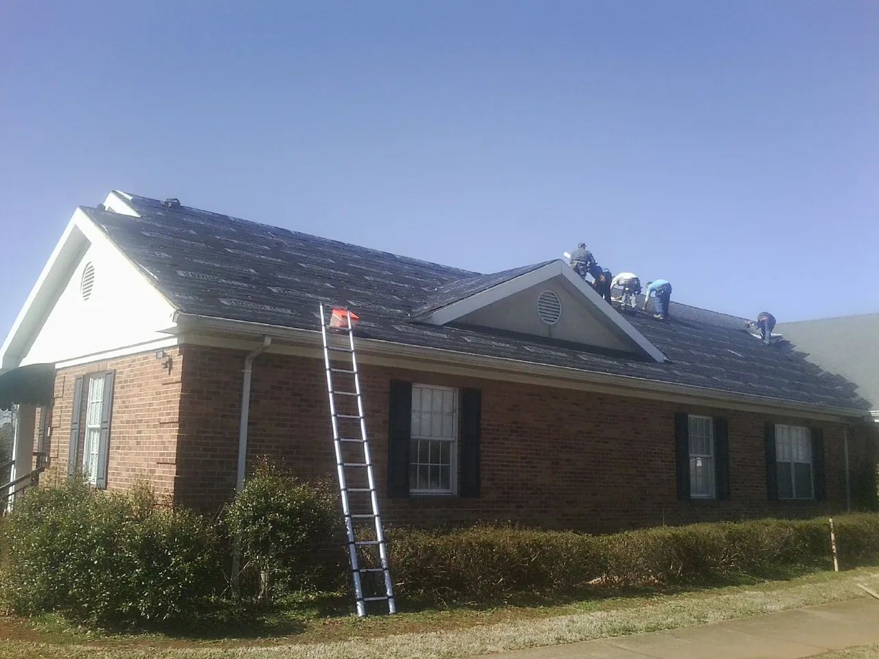 Crew tearing shingles off a brick ranch roof with a ladder set — Metal Roof Repair work by our crew in Crete