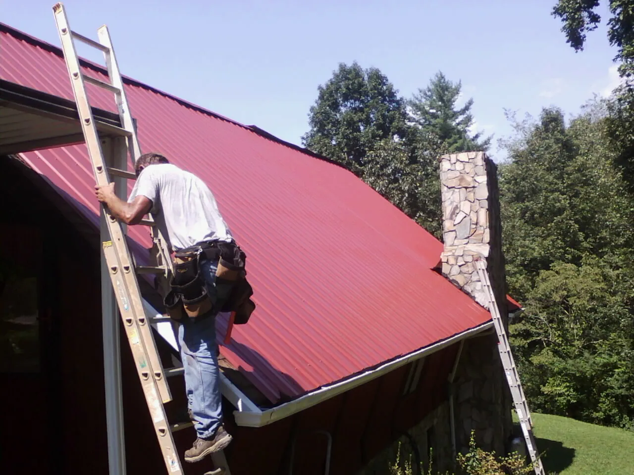 Roofer on a ladder beside a red metal roof and stone chimney — Soffit & Fascia Repair by Roofing Brothers in Crete
